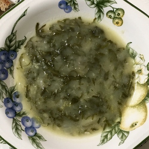 La foto muestra el caldo verde servido en el plato, realtan grumos formados por la clo gallega, previamente cortada en tiras finas, sobre un fondo verde claro.