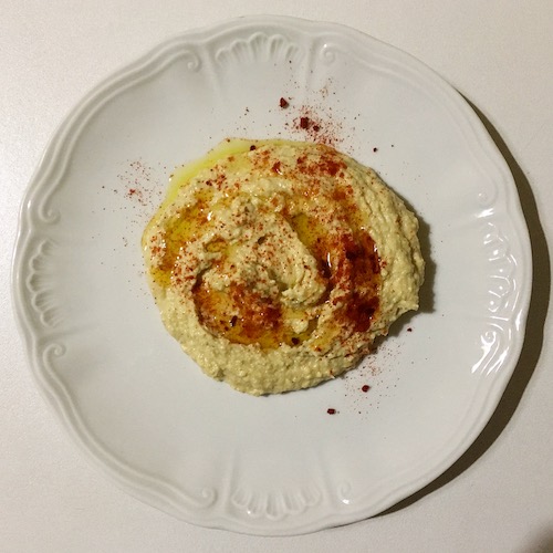 La foto muestra un plato de hummus confeccionado con garbanzos cocidos, pasta de tahina, aceite, ajos y zumo de limón. Servido con pimentón espolvoreado.